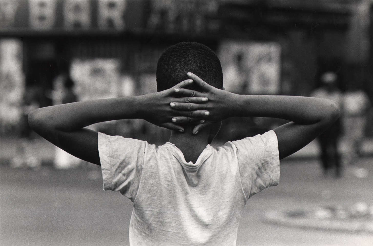 23. Beuford Smith, Untitled, ​c. 1970. Upper body of a boy photographed from behind. He wears a white t-shirt and has hands clasped behind his head.