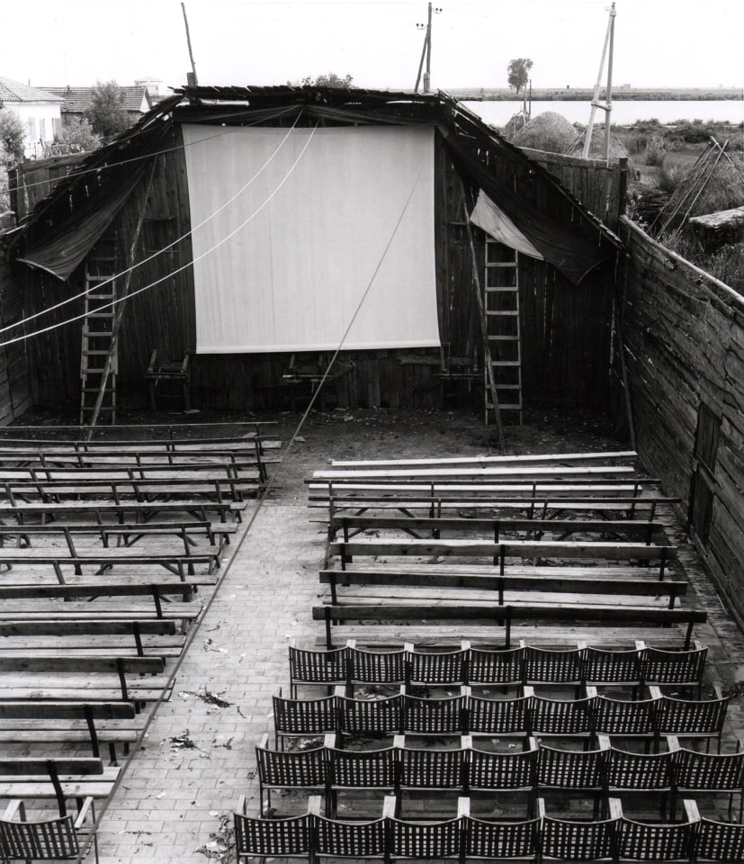 Pietro Donzelli, Cinema in Pila, Veneto, ​1954. Outdoor movie screen with rows of seats and benches in front of it. Ladders are propped up on either side of the screen.