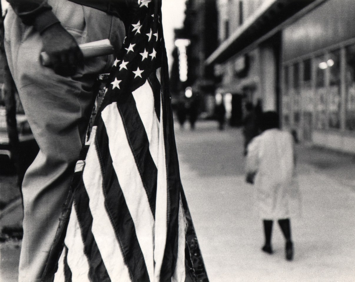 08. Beuford Smith, Street Speaker, ​1968. A man's lower body fills the left of the frame with an American flat alongside it; pedestrians pass on the sidewalk to the right.