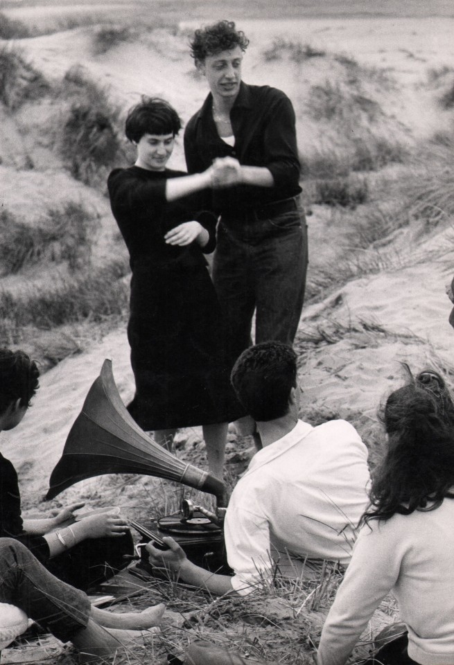 Gianni Berengo-Gardin, Venezia - Il Lido, c. 1958. A group of young adults listens to a record player on the beach. A couple in black is dancing, while the other people are seated in the sand.