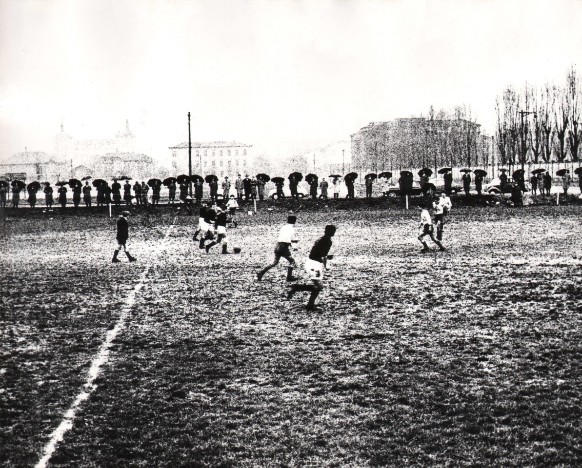Mario Carrieri, Milano, ​c. 1958. A group of young men play soccer in front of a long row of onlookers holding umbrellas.