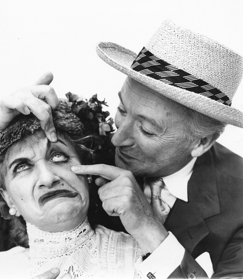 Richard Avedon, Cecil Beaton &amp; Hermione Gingold, ​1956. A suited man stands, smiling, with fingers on the face of a woman with dramatic makeup. He pulls her left eye open and she makes an exaggerated frown.