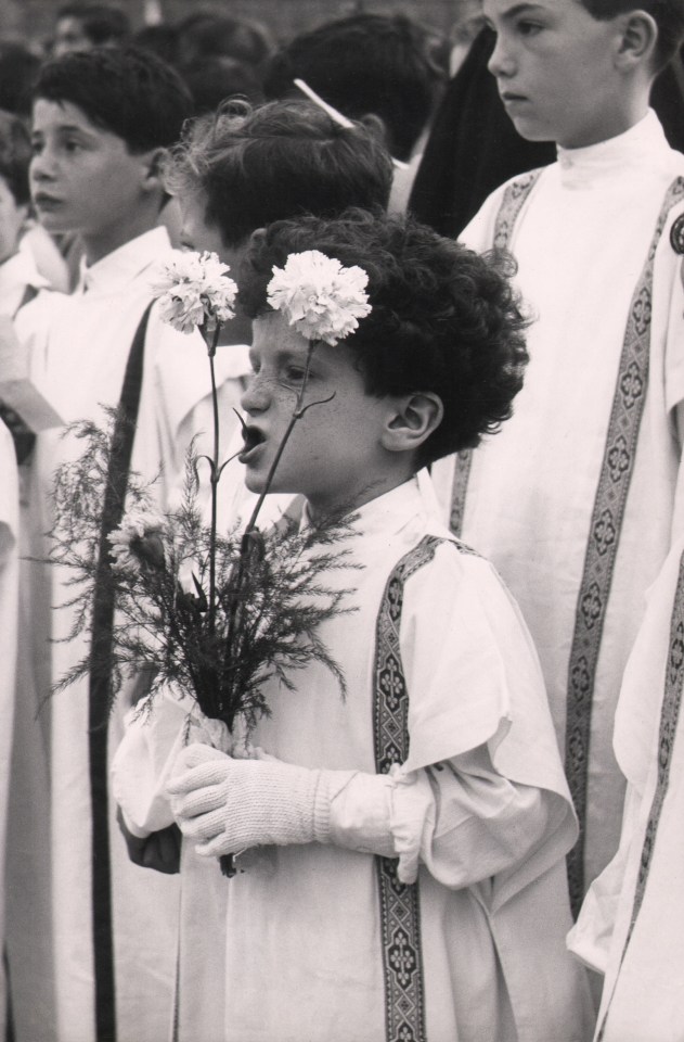 Gianni Berengo-Gardin, Venezia, processione del Corpus Domini in piazza San Marco, ​1958. A young boy among many in white robes chews on the stem of a flower he holds in front of his face.