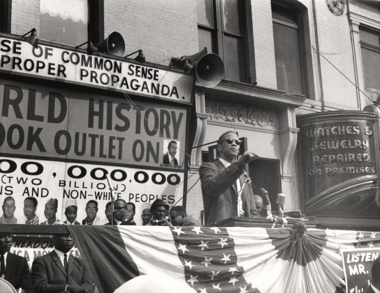 16. Beuford Smith, Malcolm X, Harlem, ​1964. Subject stands at a podium outdoors, speaking into microphones.