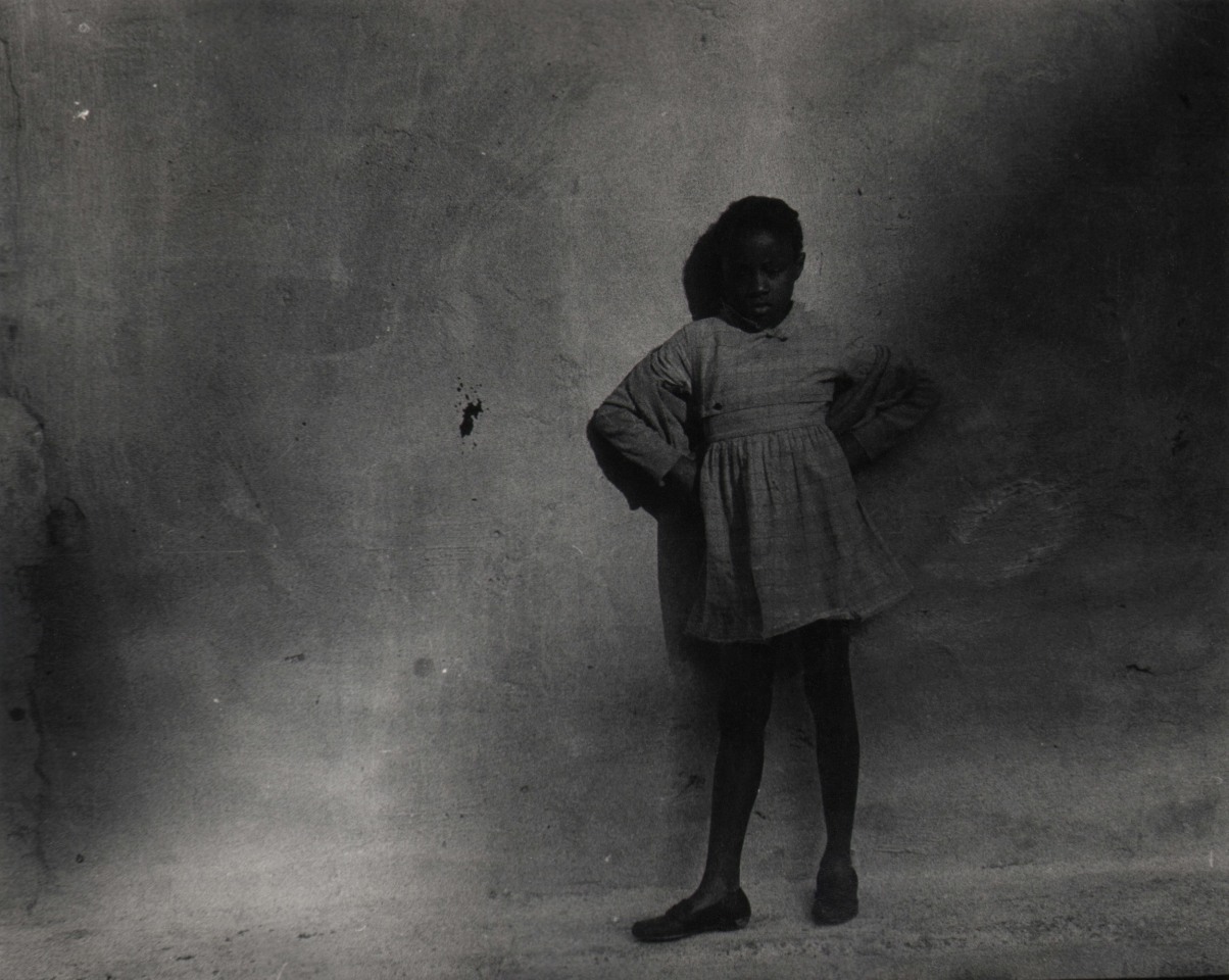 19. Beuford Smith, Little Girl in Park, ​1968. Dark exposure of a young girl standing against a concrete wall with hands on her hips.