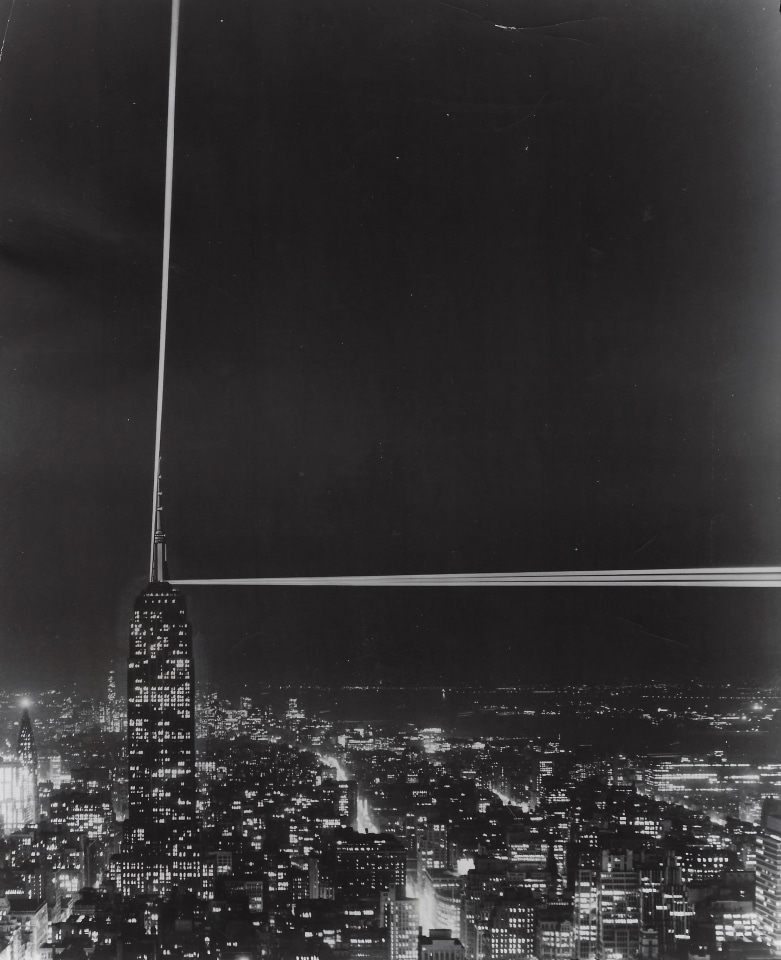 08. Anonymous, Patterns in Light: the Empire State's controversial beacons, 1956. Distant, head-on nighttime view of the Empire State Building (lower left of frame), with two beams of light streaming up and to the right.