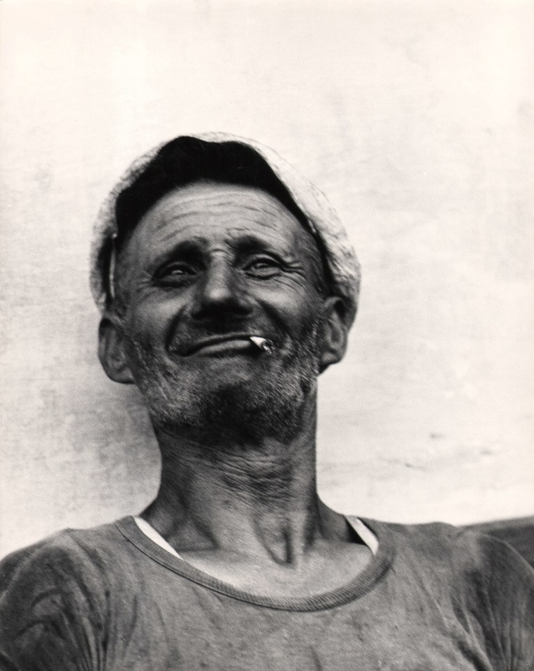 Carlo Cosulich, Operaio Italiano, 1959. A smiling man, blurred with motion, leans against a white wall, holding a cigarette between his lips.