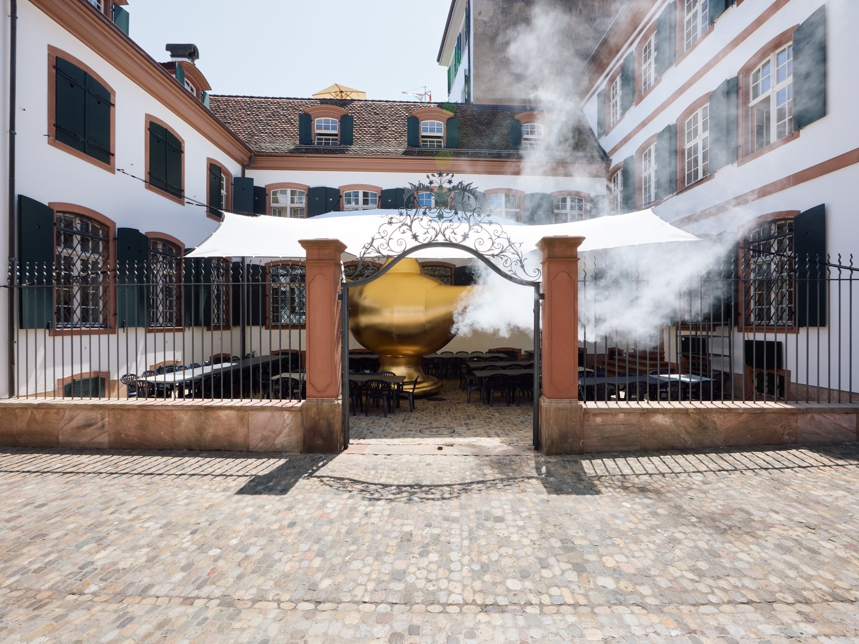 A large scale inflatable of a gold magic lamp with smoke coming out of the spout. The sculpture is installed in the center of a courtyard in Basel, Switzerland.