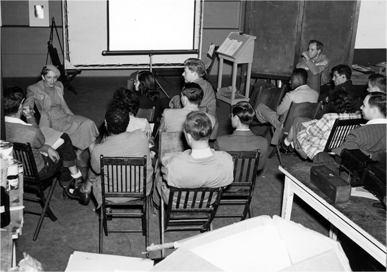 Instructor Dorothea Lange lecturing students in Studio 18; photography department chair Minor White looks on, back right, 1947.