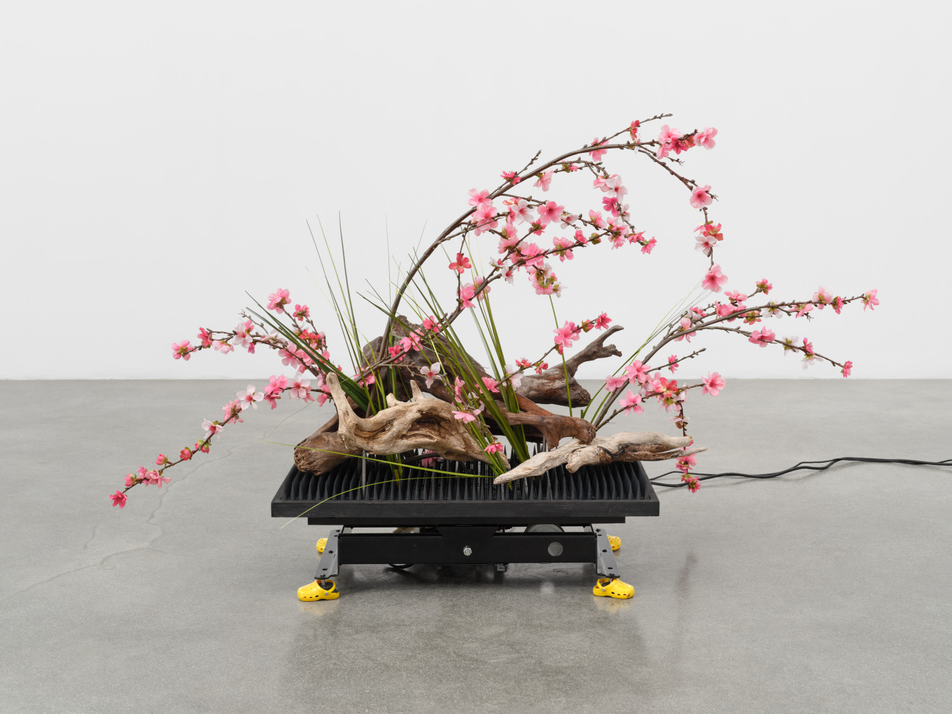 A sculpture by Rachel Youn titled &quot;Arrangement,&quot; 2023, featuring a vibration platform machine, steel, wood, plastic spikes, artificial flowers, driftwood, dog shoes, arduino, and serro motor