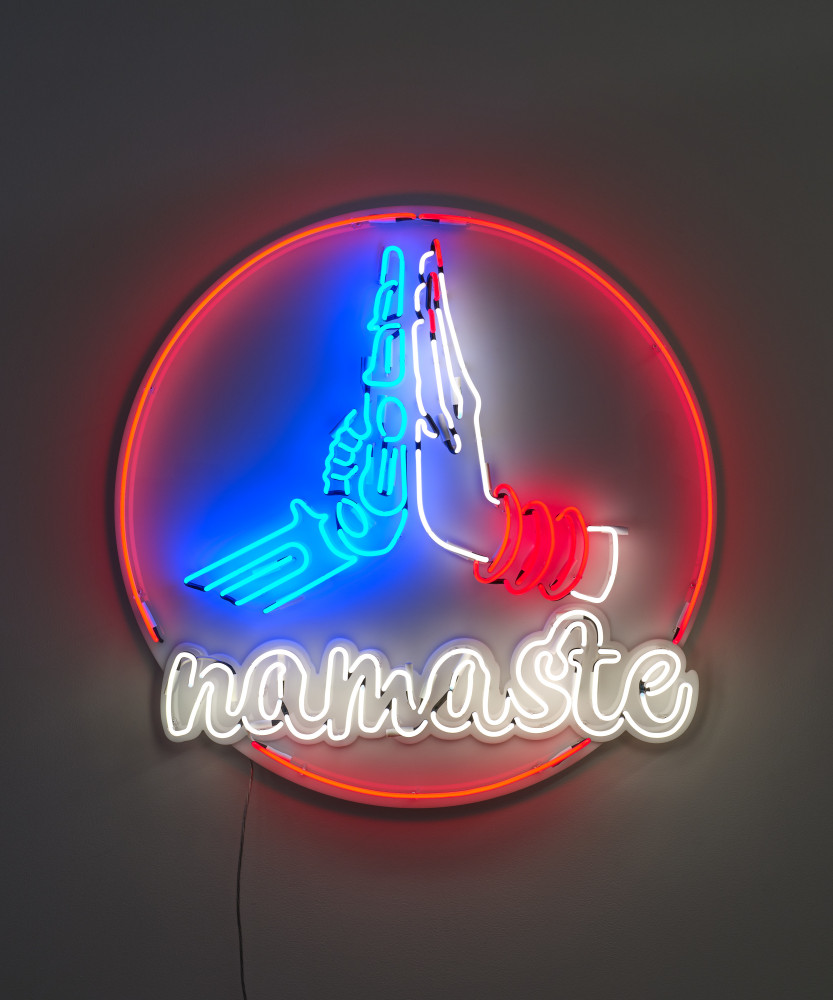 A red white and blue neon sculpture of a robotic hand pressed in prayer position against a female human hand with text that reads &quot;namaste&quot; encircled in a read neon circle.