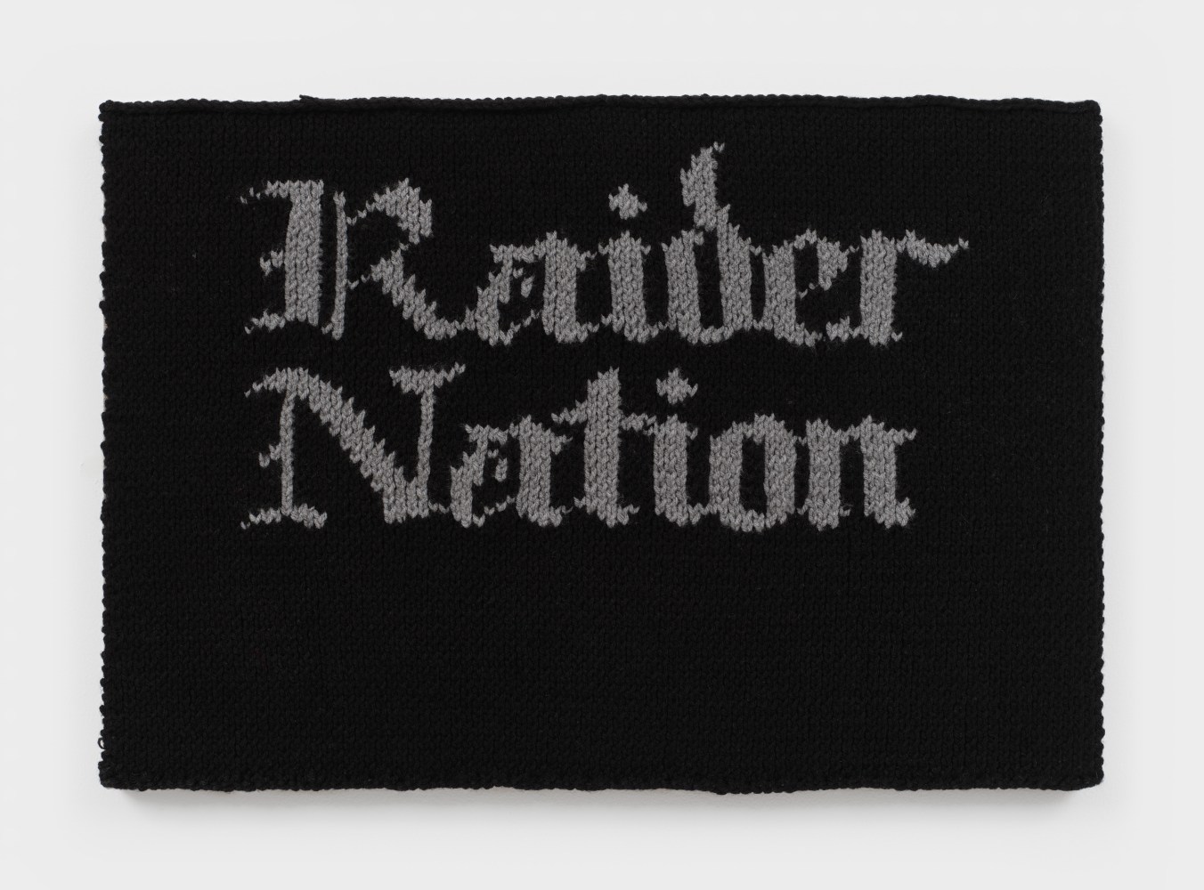 A knit artwork with the words &quot;Raider Nation&quot; in grey against a black background, sewn onto linen and stretched.