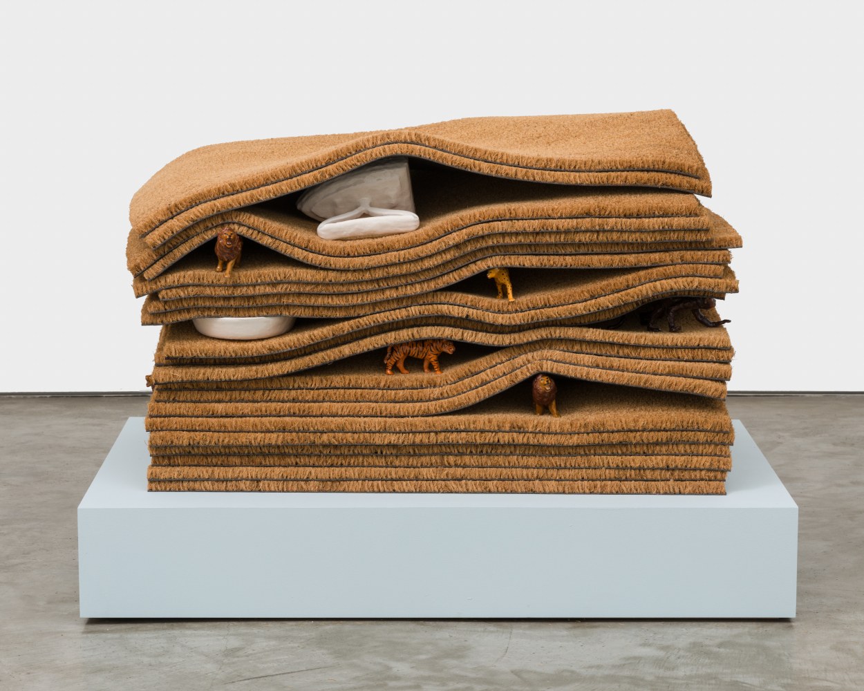 A sculpture with 20 layered doormats with ceramic animal figurines and ceramic irons peaking out from between the layers.
