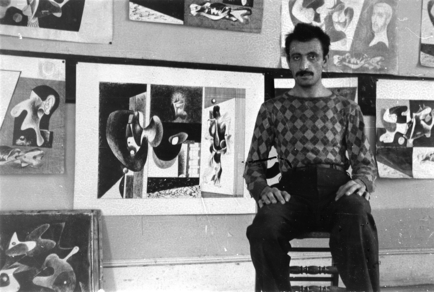 Black-and-white photograph of a man in a patterned sweater seated before a wall on which eight abstract ink drawings are hung