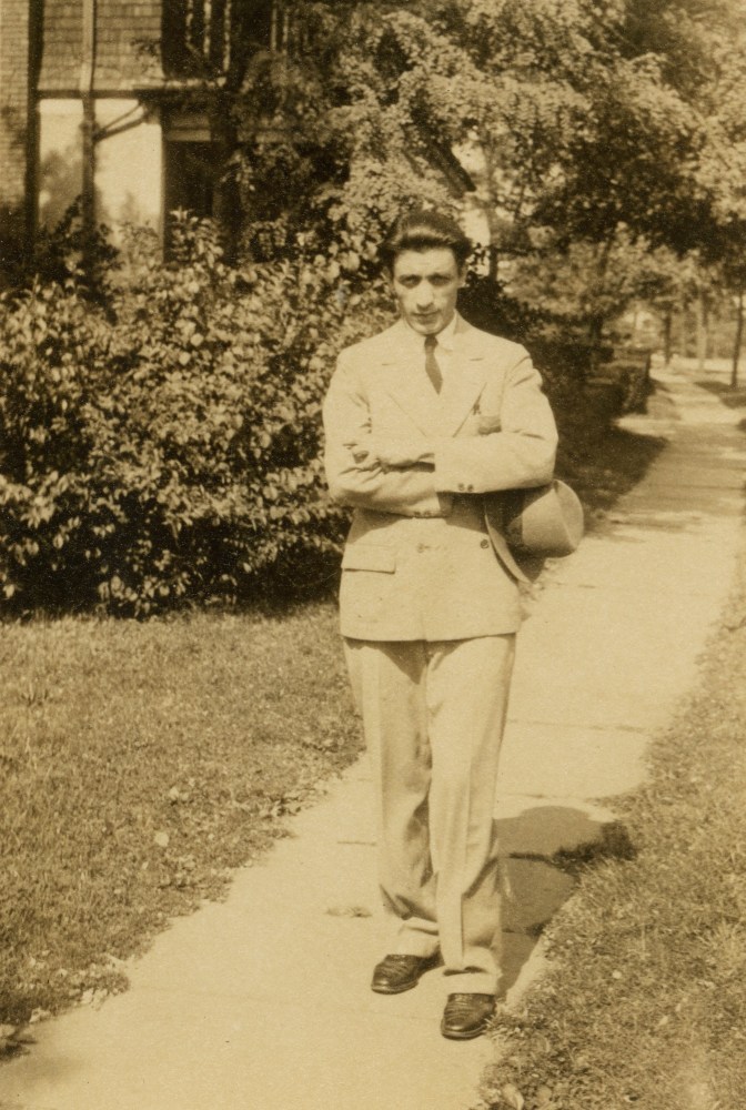Sepia photograph of a male dressed in a suit standing on the sidewalk of a residential city street