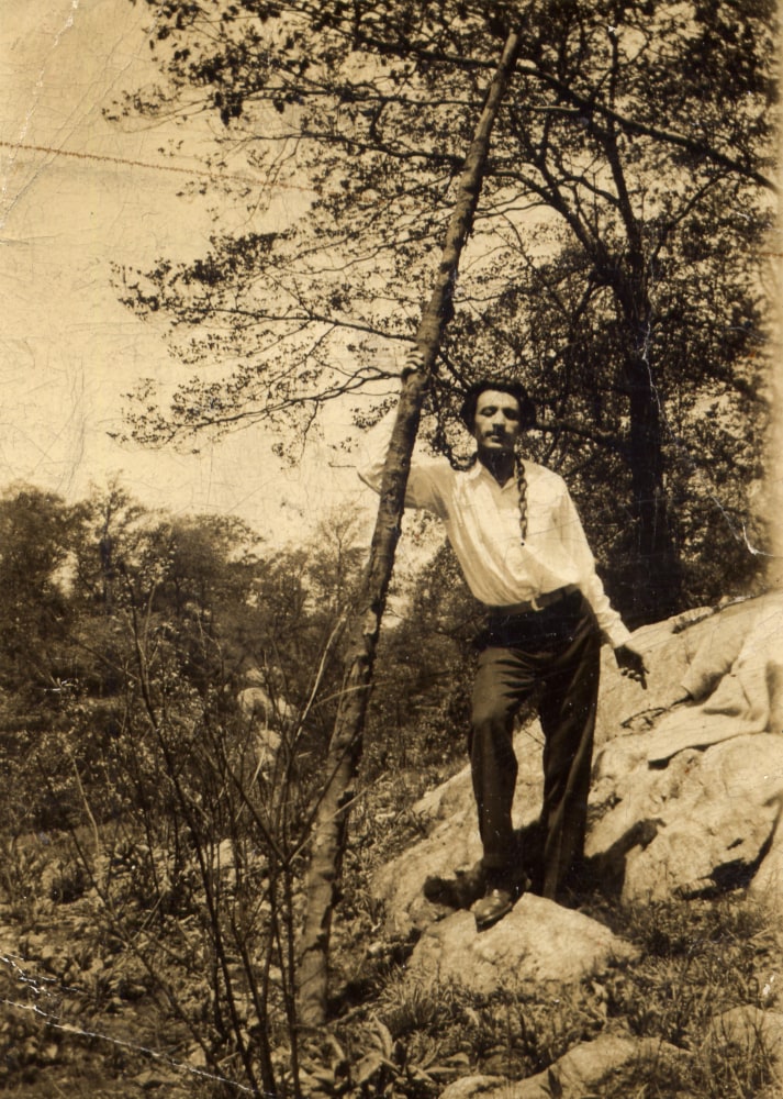 Standing man with black hair in black pants, a white shirt, and a chain around his neck posing against a thin tree in a rocky park