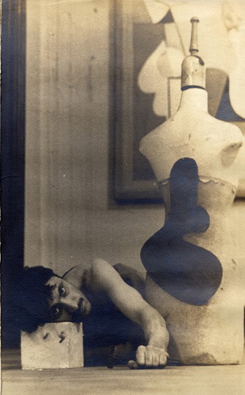Young man reclining in a studio, with his head resting on a block, near a mannequin with an abstract shape and behind him a detail of an abstract painting hanging on the wall
