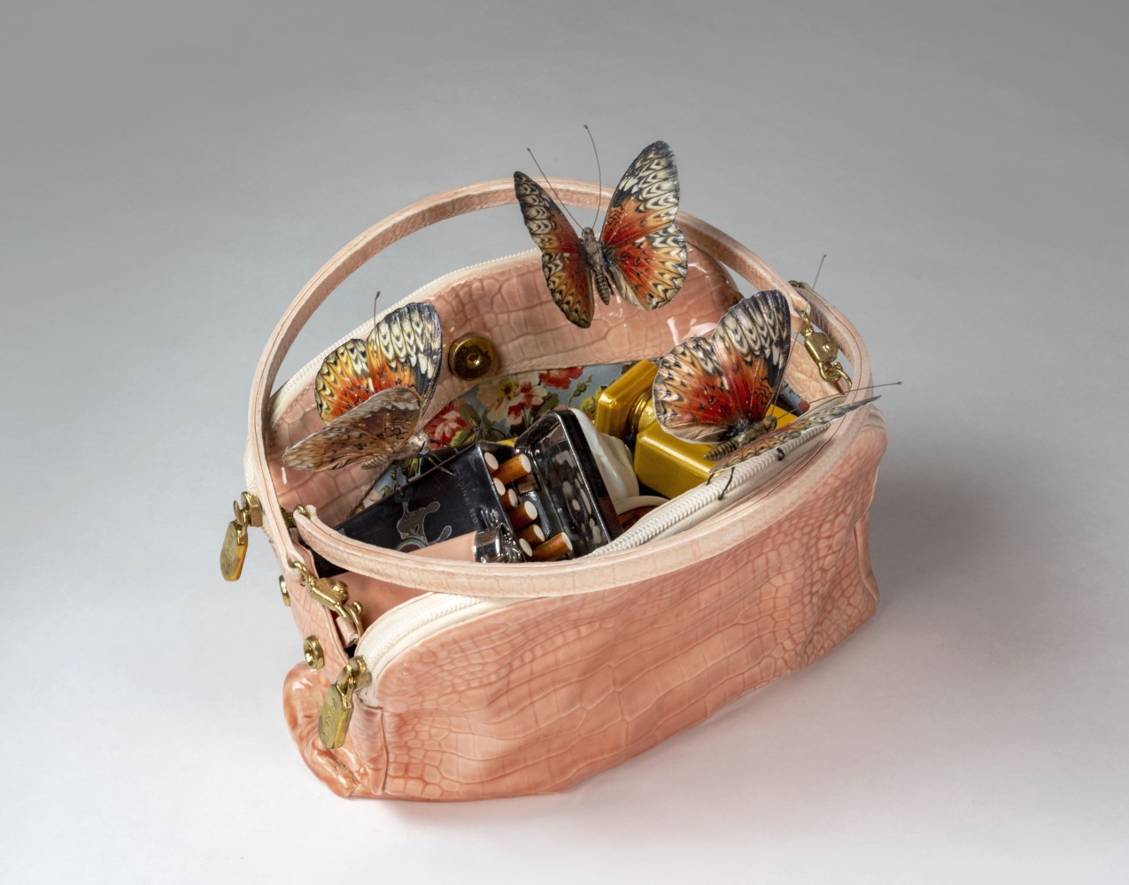 A pink ceramic handbag with three butterflies perched atop the bag's open compartment, revealing the bag's floral interior lining, a metal tin of cigarettes, perfume, and a matching pink lighter