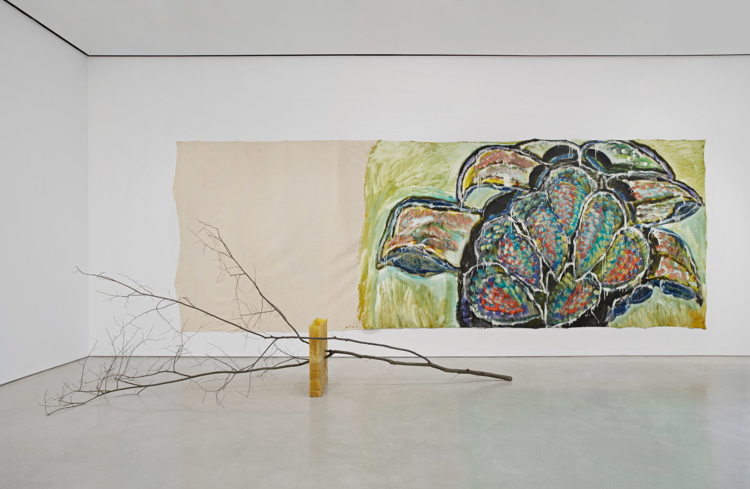 a large branch on its side supported by a wax block while a large-scale painting of foliage hangs on the wall behind it