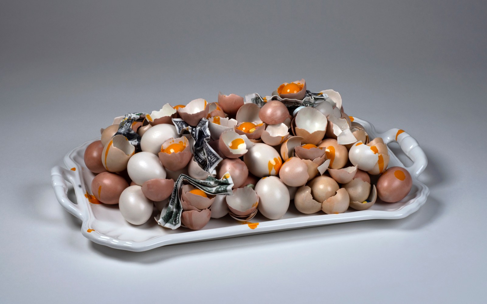 A ceramic tray with two handles holding a number of eggs and broken eggshells with crumpled dollar bills and runny egg yolks