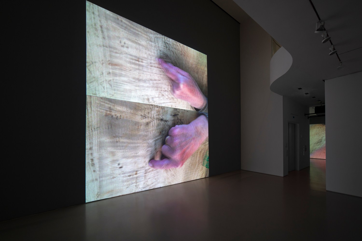 gallery installation view of two stacked projections of hands with index fingers extended drawing patterns on a marked wooden surface