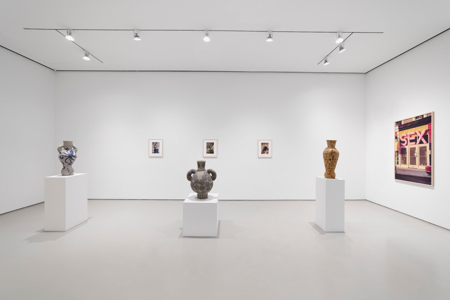 3 ceramic sculptures on pedestals with 3 photographs hanging behind on a white wall and one larger photograph on the right adjacent wall