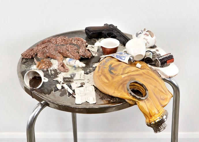 stainless steel and ceramic table holding an octopus, spilled espresso cups, a gun, batteries and a gas mask