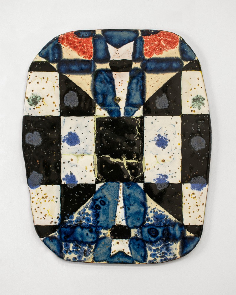 Oval ceramic wall piece with a patchwork of black, cream, red, and deep cobalt-blue glazed sections arranged in a quilt-like grid. The surface features speckled and bubbled textures, with areas of mottled blue resembling ink blots or water stains.