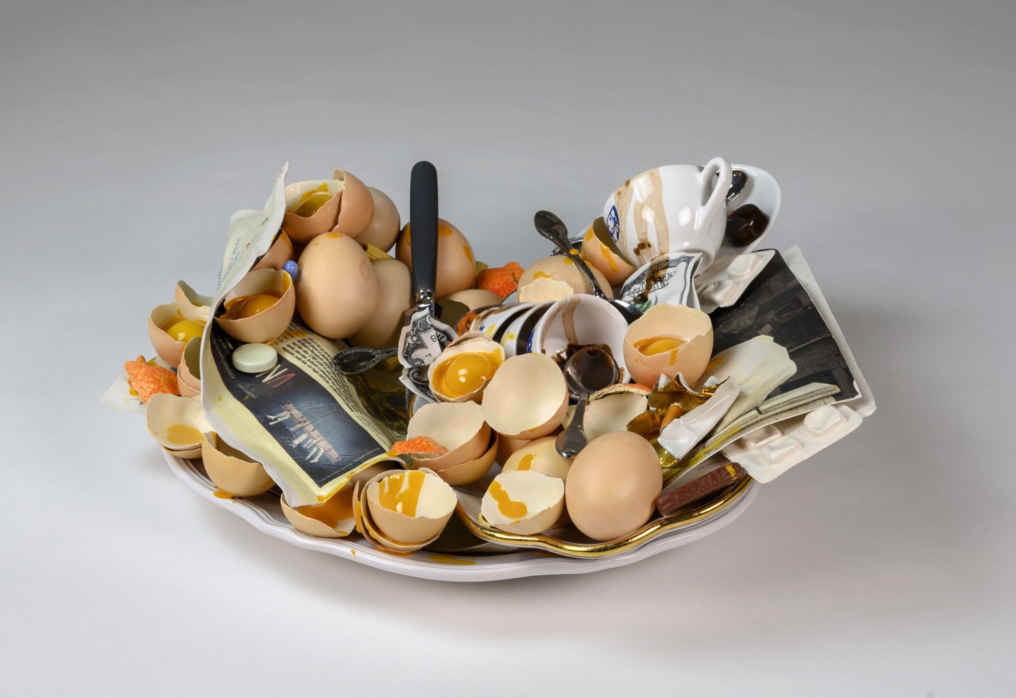 A cermic tray holding a number of broken eggshells and their yolks, piled messily amongst a page from a publication, crumpled dollar bills, and coffee-stained espresso cups and spoons