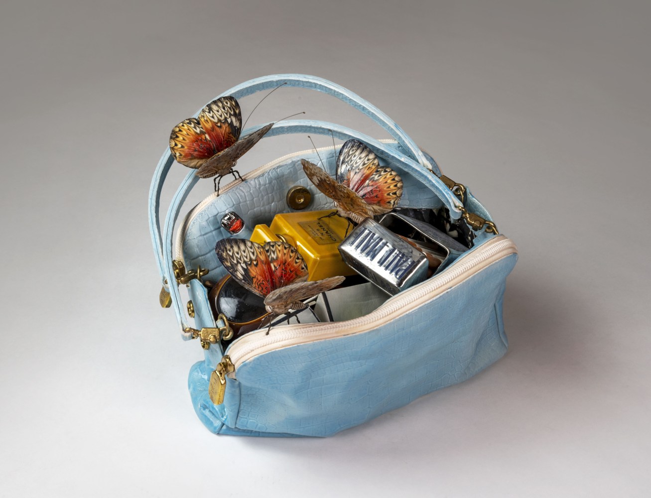 A blue ceramic handbag with three butterflies perched atop the bag's open compartment, revealing perfume, cigarettes, and a lighter