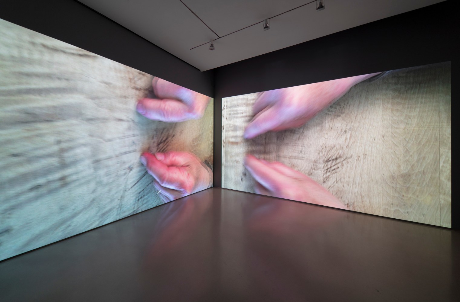 installation view of two video projections showing large hands over a wooden table