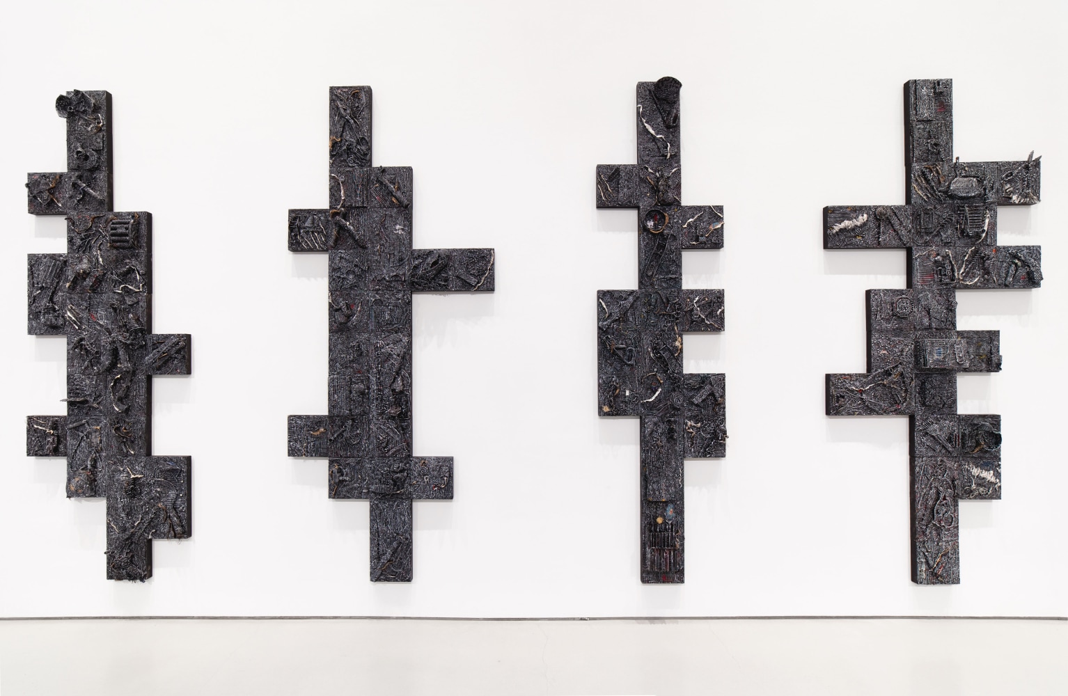 Four vertical, dark, textured, and geometric artworks hang on white gallery wall.