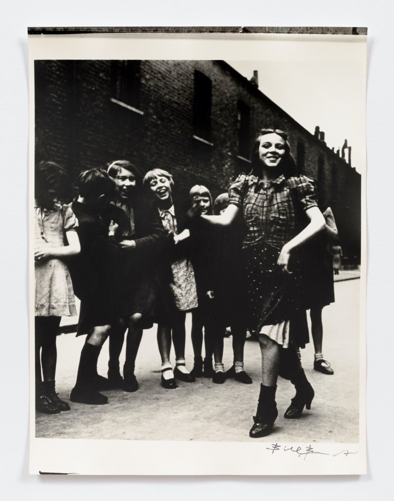 Black and white photographic portrait of girls dancing the Lambeth Walk in the 1930s in front of a dark brick building.