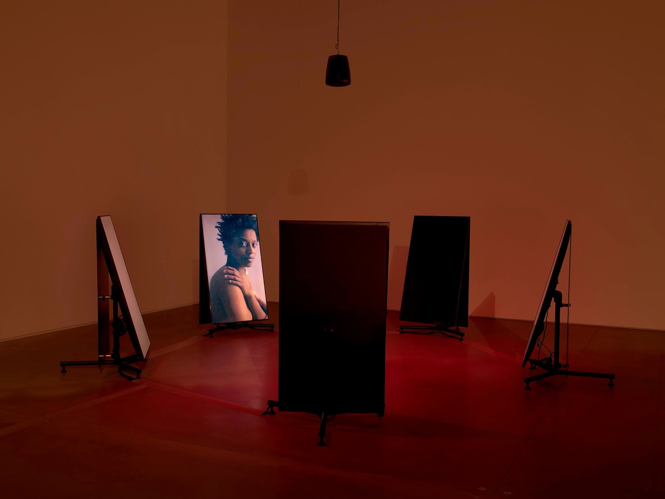 Photo of five video monitors in dim red lighting which feature an image of a woman