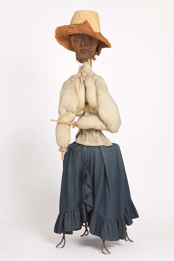 A sculpture made out of Hogmawg, fabric, and mixed media that depicts a women wearing a hat, white blouse, and denim skirt.