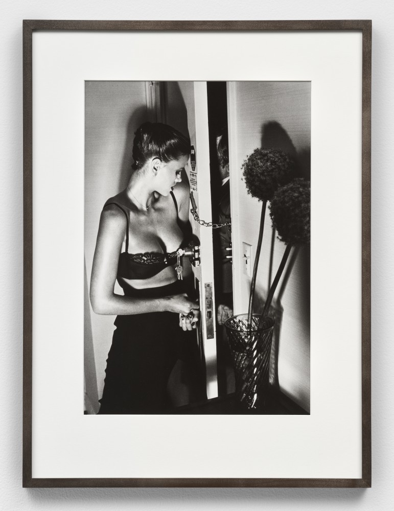Black and white photographic print by Helmut Newton of a woman standing near a chain-locked door with a man outside of it