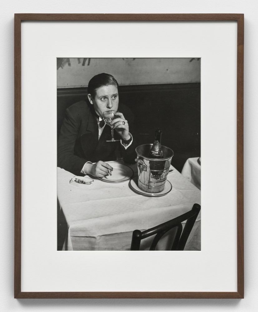 A black and white photographic print by Brassai depicting a woman dressed as a man with a wine glass in hand sitting a white top table