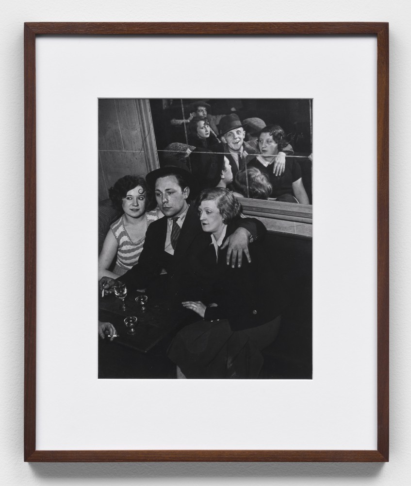 A black and white photographic print by Brassai of three people sitting down with glasses and the reflection of a group of people in the mirror