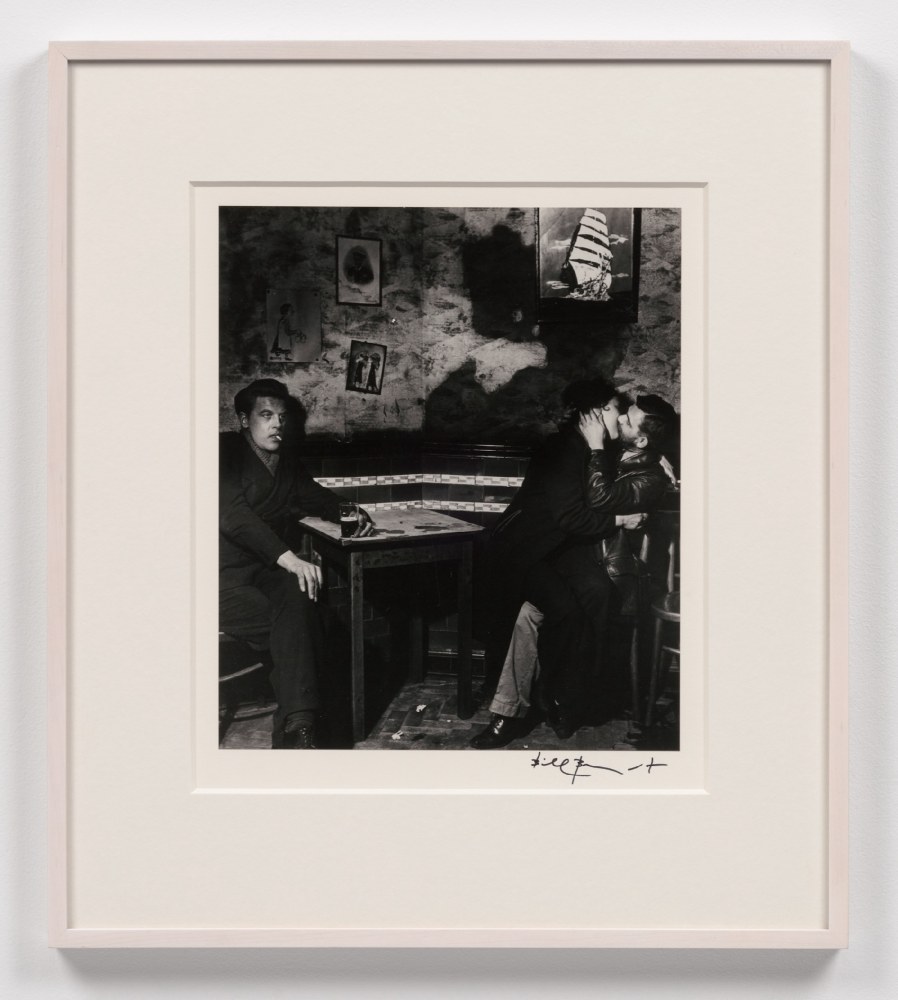 A black and white Bill Brandt photographic print depicting a couple kissing and a separate person across the table with a cigarette and drink