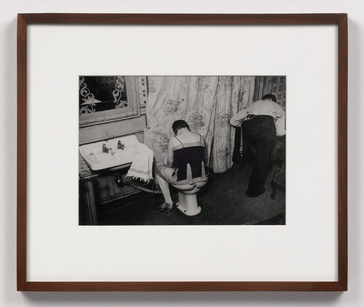 A black and white photographic print by Brassai of a person sitting on a toilet in a brothel with another person to their right