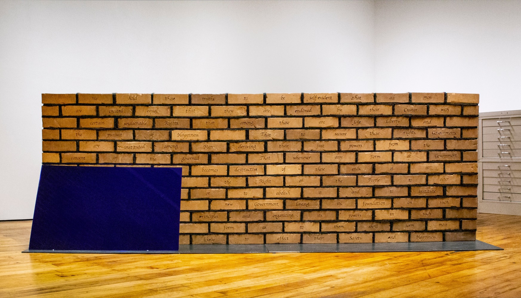 Sonya Clark
Edifice and Mortar, 2018
Hand stamped bricks, human hair, and glass, steel base
39 x 72 x 15 in. (99.9 x 182.9 x 38.1 cm)
Clar-1034-C

Sonya Clark describes&amp;nbsp;Edifice and Mortar&amp;nbsp;as a wall, a flag, and a document that asks us to consider a fundamental question: who laid the&amp;nbsp;foundations of the United States of America?

&amp;nbsp;

Each brick is hand-stamped with a traditional maker&amp;rsquo;s mark and a word. Together the recto forms an excerpt from the Declaration of&amp;nbsp;Independence. The lines of text are interspersed with mortar made from African American hair gathered from Richmond salons&amp;mdash;the hair of&amp;nbsp;people whose ancestors might have been legally enslaved and whose life, liberty, and the pursuit of happiness were cut out of Jefferson&amp;rsquo;s 1776&amp;nbsp;Declaration. The viscerally charged pointing is meant to represent &amp;ldquo;black people who are at once held under the weight of the system, but who&amp;nbsp;are also holding this country together.&amp;rdquo;

&amp;nbsp;

The brick stamps display the word&amp;nbsp;Schiavo, which is the origin of the commonly used Italian greeting Ciao. Translated into English, the word&amp;nbsp;Schiavo means SLAVE.

&amp;nbsp;

The blue panel is intentionally placed at an angle to reflect the viewer, making each of us a part of the work.

&amp;nbsp;

Edifice and Mortar&amp;nbsp;extends Clark&amp;rsquo;s ongoing material and conceptual reworking of icons of America&amp;rsquo;s racially divided past and present.