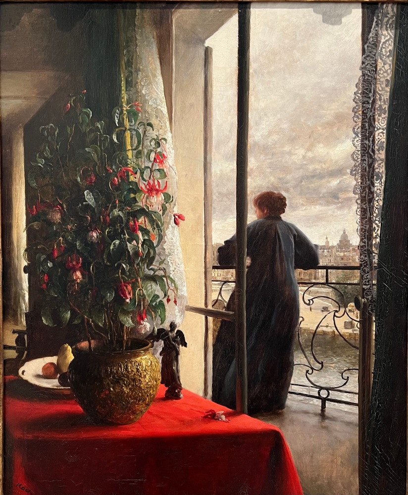 back of female figure standing on a balcony in Paris and in the foreground is a large flowering pland