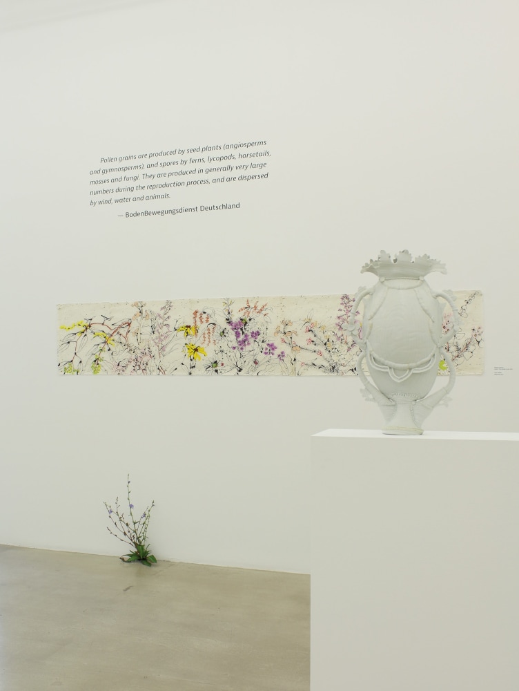 Photograph of a detail view of an exhibition with a long horizontal drawing hung on a white wall, with a sculpture on the ground and a sculpture on a pedestal in front