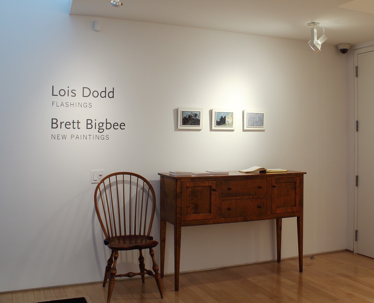 Photograph showing a front view of an exhibition space, with three small paintings hanging on a white wall above a table and chair