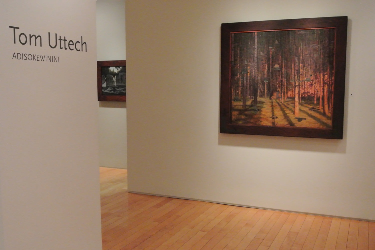 Photograph showing a view of an exhibition space, with one framed painting hanging on a wall to the right and wall text that reads &quot;Tom Uttech: Adisokewinini&quot; on the wall to the left