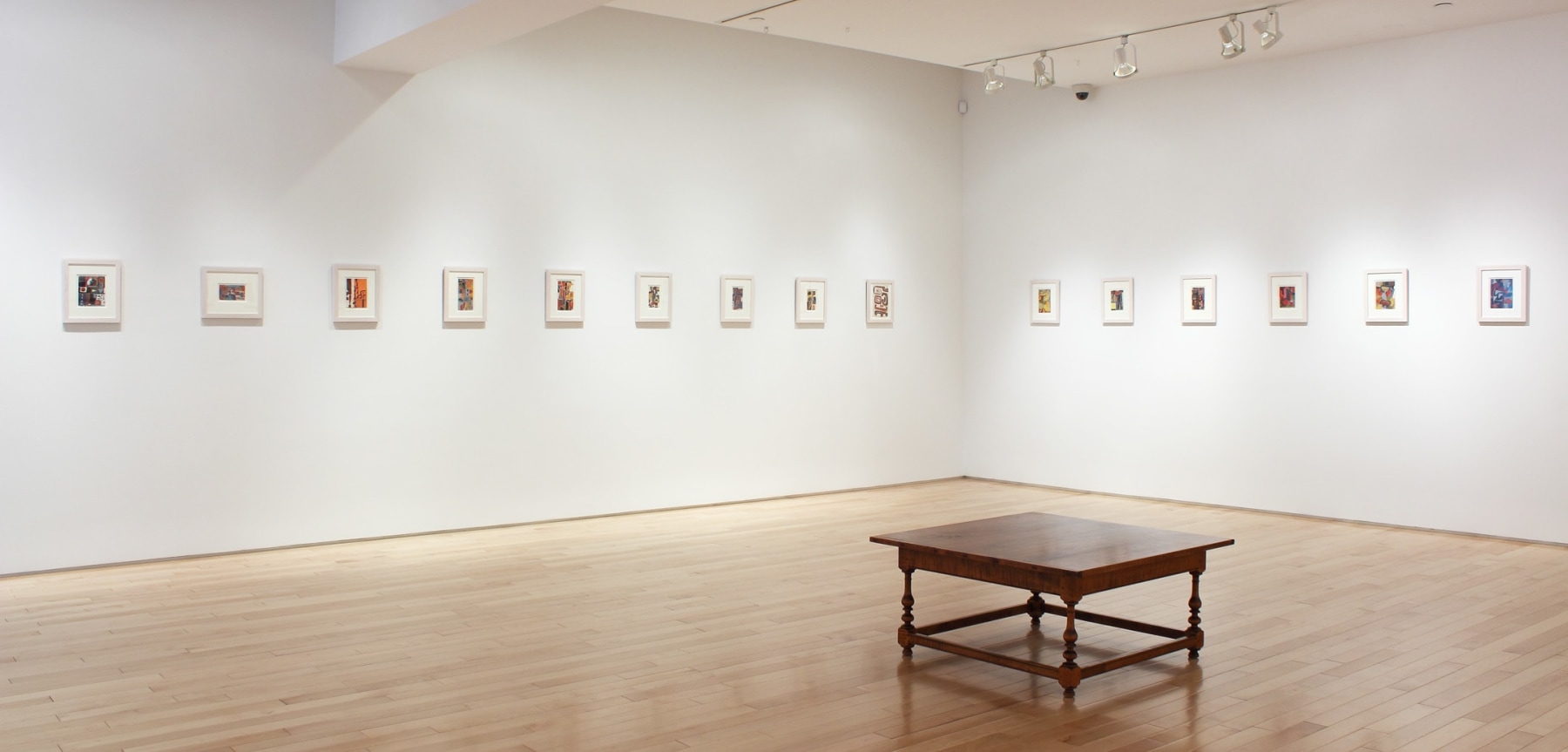Photograph showing a corner view of an exhibition space, with fifteen small framed paintings hanging on white walls