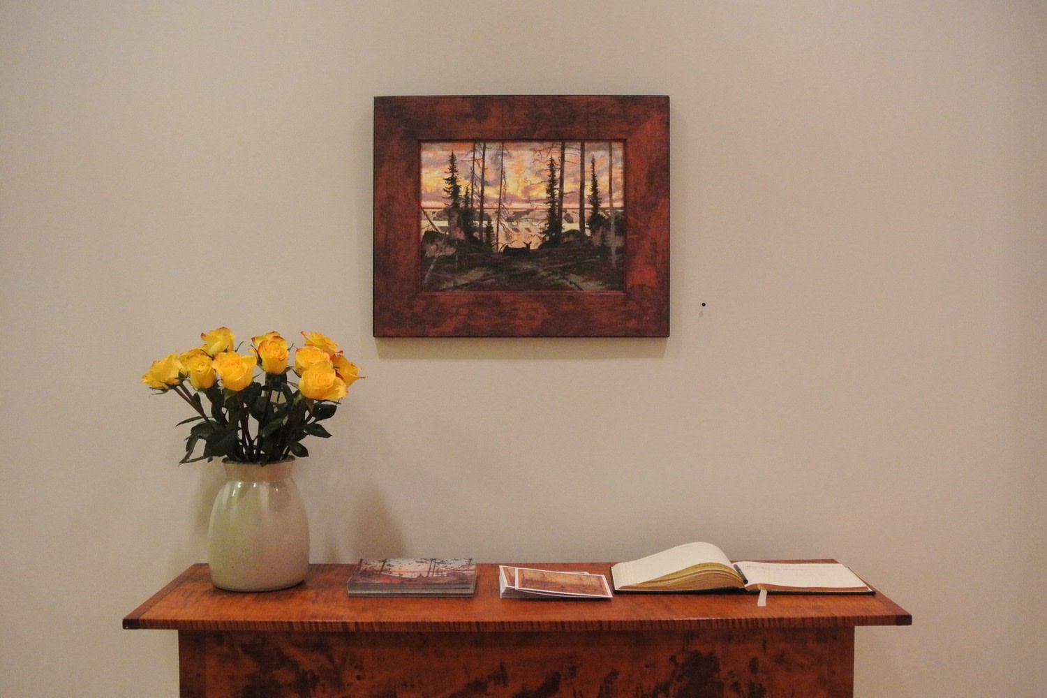 Photograph showing a view of an exhibition space, with a small framed painting hanging over a table with a guestbook and vase of flowers on it