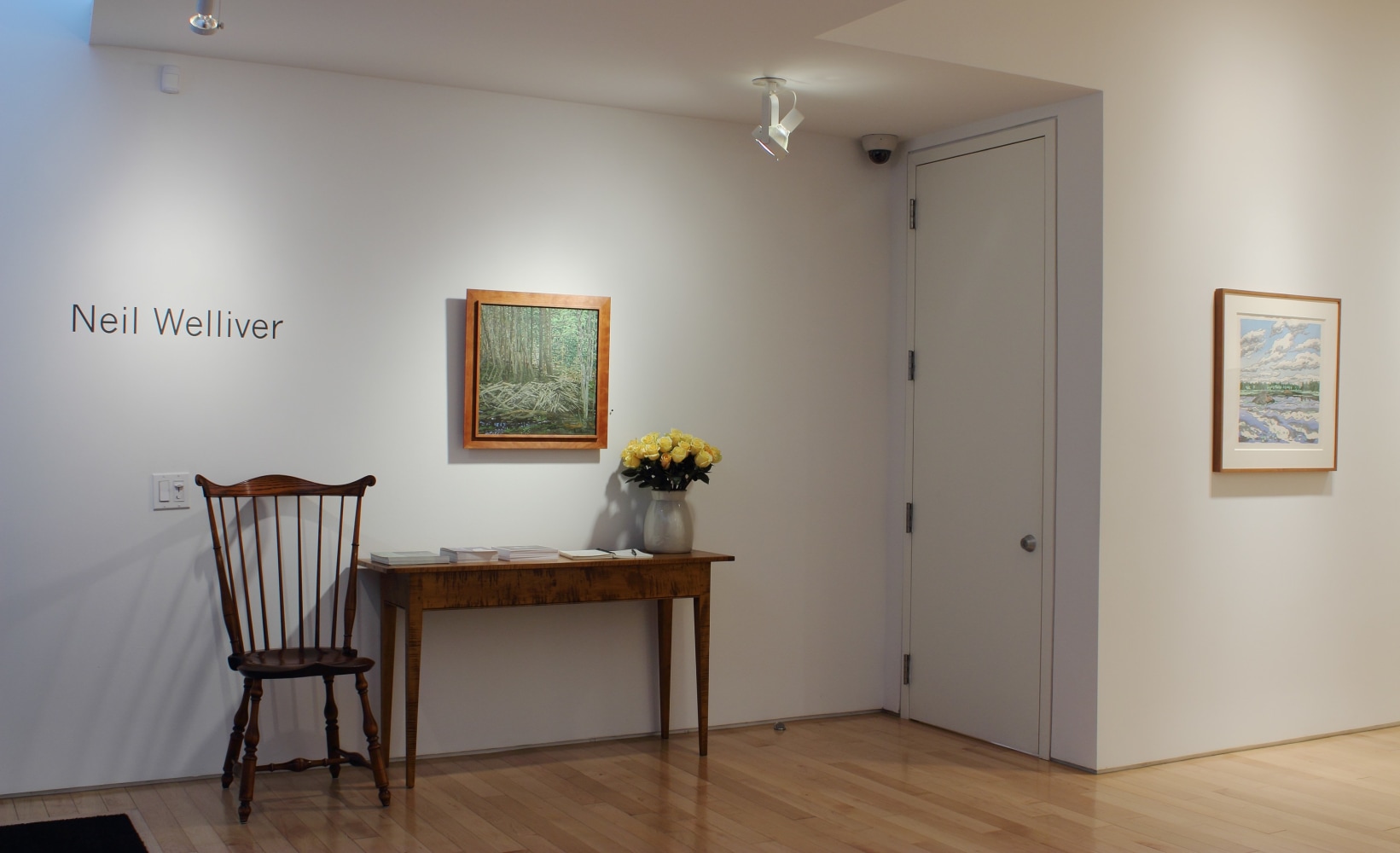 Photograph showing a view of an exhibition space, with one painting hanging on a white wall over a table a chair