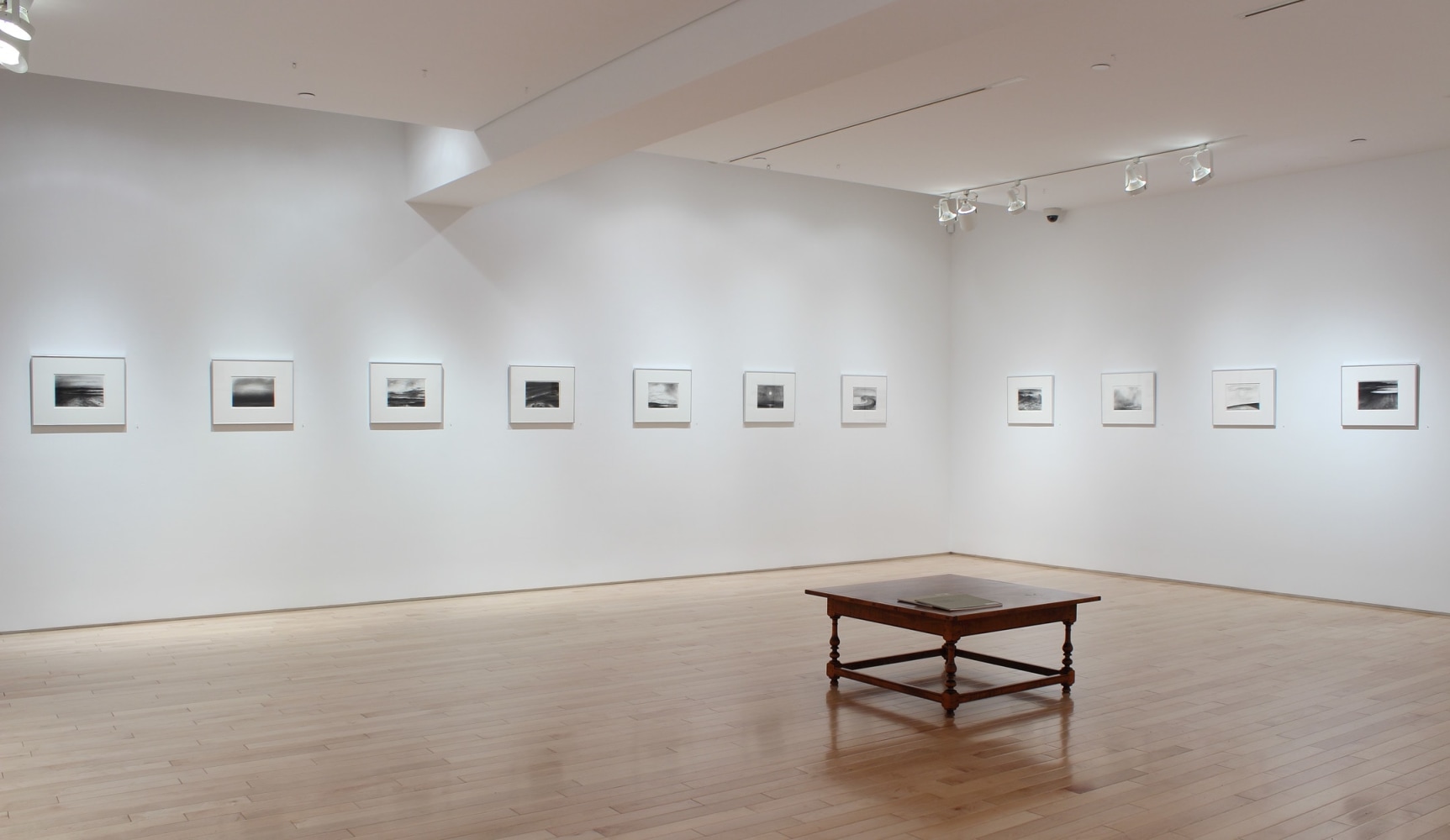 Photograph showing a corner view of an exhibition space, with thirteen drawings hanging on white walls