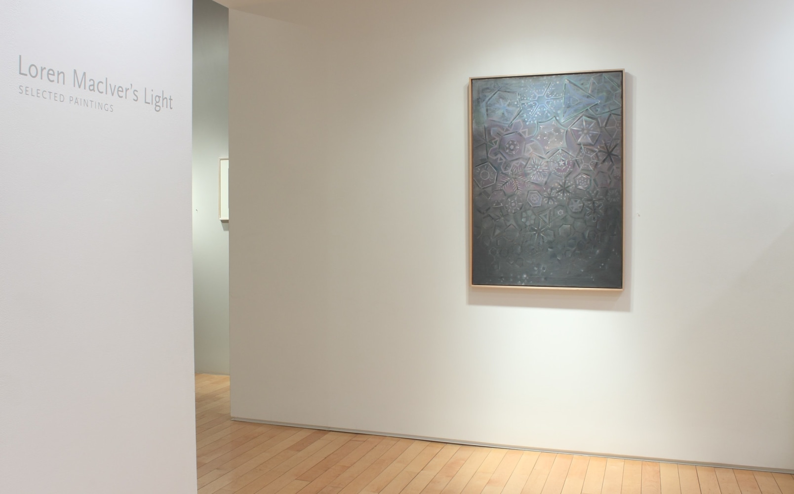 Photograph showing a view of an exhibition space, with one painting hanging on a wall and text that reads &quot;Loren MacIver's Light: Selected Paintings&quot; on another wall to the left